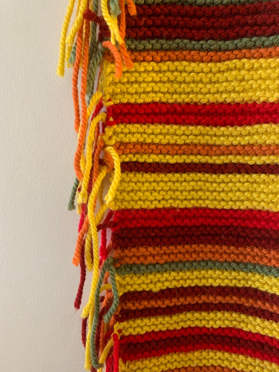 A close-up of a knitted scarf shows stripes in orange, yellow, green and brown. Each strip represents a week of the year, and each color represents a different concentration of the carcinogen chloroplene.